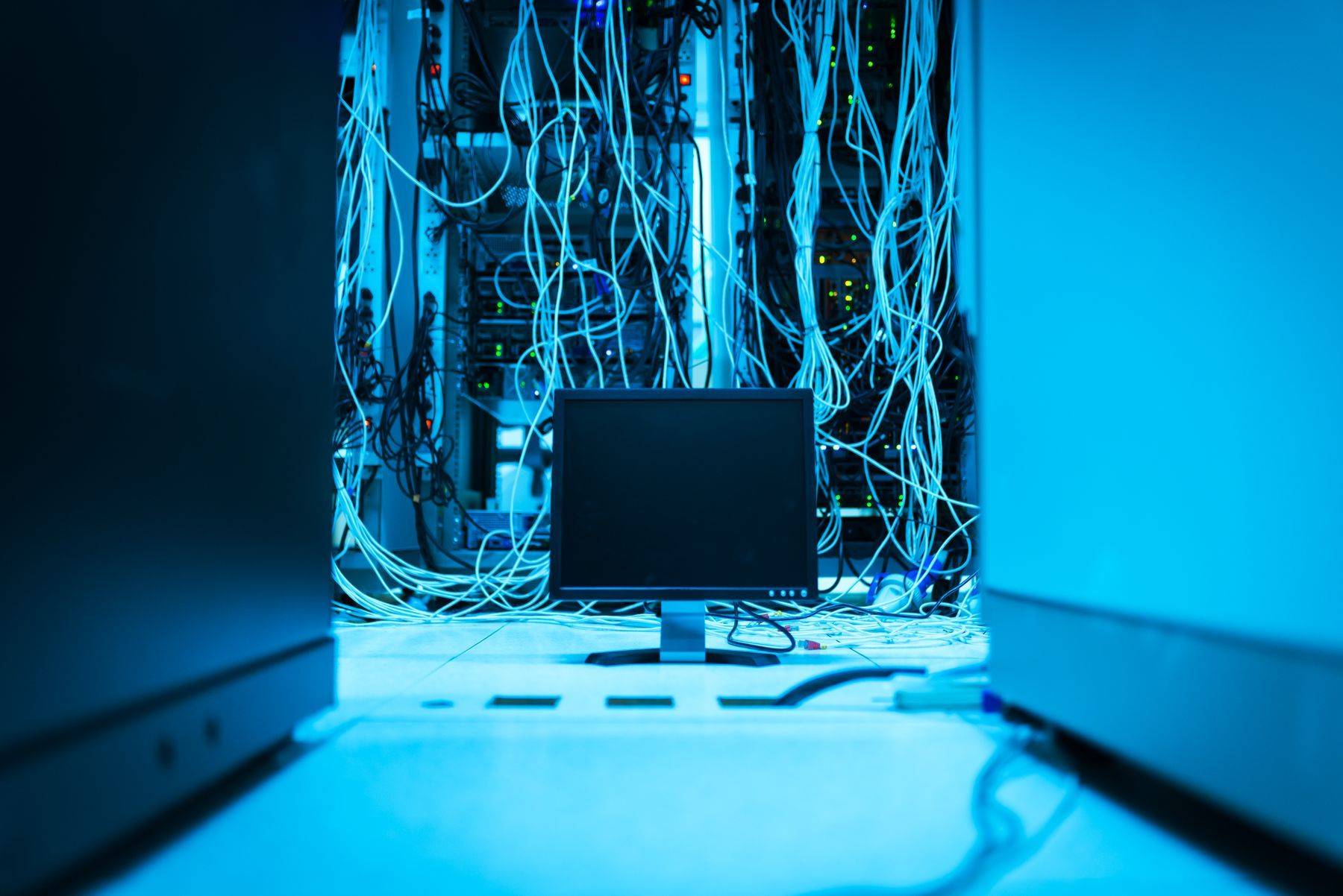 Cabluri de rețea în spatele unui rack și un monitor, într-o cameră cu lumină albastră